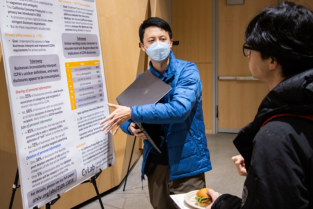 An asian male in a mask holding a laptop presented to another young man his research poster