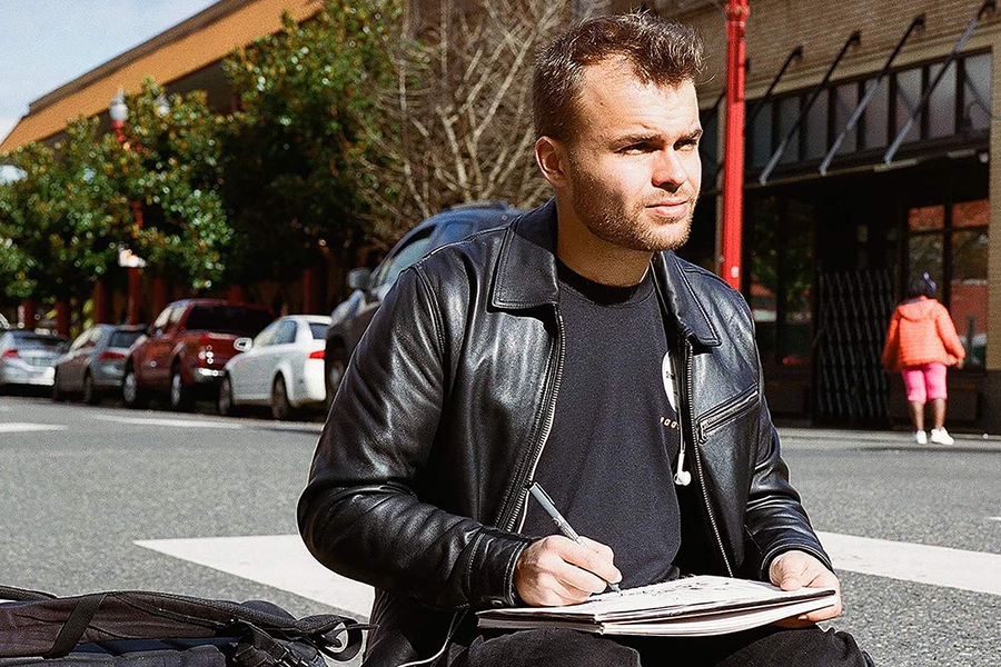 David widder wearing a leather jacket, black tee, and combat boots, sitting on a street corner sketching in his notebook