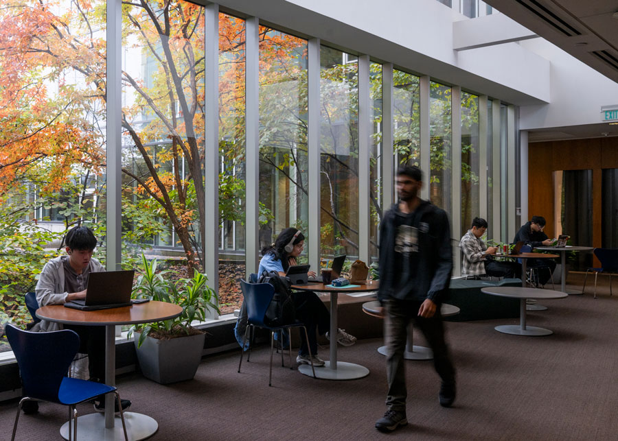 A slightly blurred man walks through a public corridor where students study at tables against a background of large windows.